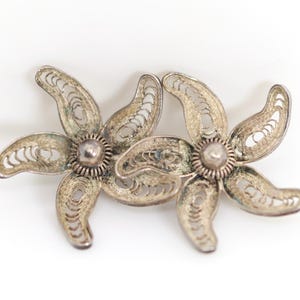Könnte beinhalten: Ein Paar silberfarbene Metallohrringe mit einem fünfblättrigen Blumendesign. Jeder Ohrring ist mit komplizierten Drahtfiligranmustern in den Blütenblättern und einer kleinen, glatten, runden Metallperle in der Mitte der Blume gestaltet.
