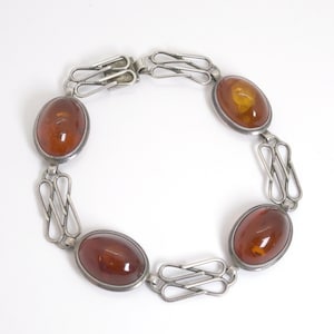 Könnte beinhalten: Ein silberfarbenes Armband mit vier ovalen, durchscheinenden bernsteinfarbenen Steinen in Metallfassungen. Die Steine sind durch durchbrochene Metallglieder mit einem geometrischen Muster verbunden und mit einem Metallverschluss versehen.