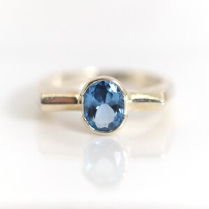 Könnte beinhalten: Ein Silberring mit einem zentralen, oval geschliffenen blauen Edelstein in einer Zargenfassung. Die Ringschiene hat eine glatte, polierte Metalloberfläche mit einem flachen, breiten Design, das sich nach hinten leicht verjüngt und klar reflektiert.