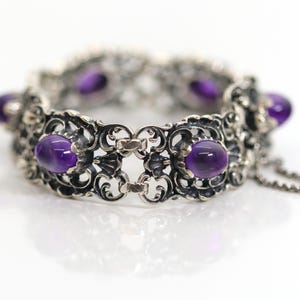 May include: A silver bracelet featuring an intricate filigree design with multiple oval-shaped, deep purple amethyst cabochons set within the metalwork. The piece includes a delicate chain link closure and exhibits a polished, reflective finish.