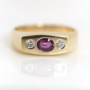 Könnte beinhalten: Ein Goldring mit einem zentralen oval geschliffenen violetten Edelstein, flankiert von zwei kleinen runden, klaren Diamanten. Die Steine sind bündig in ein glattes, poliertes Goldband mit einem flachen, rechteckigen Oberteil eingelassen.