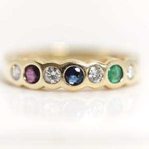 Könnte beinhalten: Ein Goldring mit sieben in einer Reihe gefassten Edelsteinen. Die Steine umfassen klare Diamanten, einen tiefblauen Saphir, einen leuchtend grünen Smaragd und einen rötlich-violetten Stein, alle in einer Fassung aus poliertem Gelbgold.