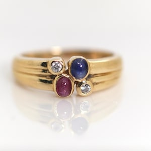 Könnte beinhalten: Ein Goldring mit einer Anordnung aus vier Steinen: einem ovalen blauen Saphir, einem ovalen roten Rubin und zwei kleinen runden, klaren Diamanten. Das Ringband ist glatt und poliert, wobei die Steine mittig auf der Vorderseite sitzen.