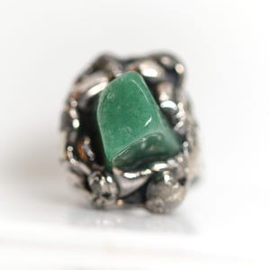 Könnte beinhalten: Ein Ring mit einem zentralen, polierten, unregelmäßigen grünen Stein, der in ein dunkles, strukturiertes Metallband gefasst ist. Das Metall hat ein raues, organisches Aussehen mit einem anthrazitfarbenen Finish, das den glatten Stein umschließt.
