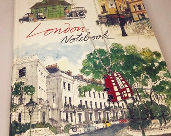 Puede incluir: Un collar plateado con un colgante de autobús de dos pisos rojo de Londres cuelga sobre un cuaderno. La portada del cuaderno presenta una ilustración de acuarela de escenas de Londres y el texto "London Notebook".