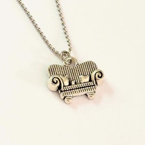 May include: Silver-tone necklace with a miniature couch pendant. The pendant has a striped back, scrolled arms, and two small pillows. The necklace chain is a delicate ball chain style.