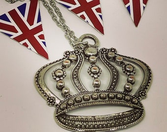 May include: A silver crown pendant necklace with a chain. The pendant is detailed with small beads and has a rounded top. The necklace is hanging against a background of three red, white, and blue fabric flags.