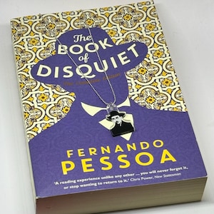 Może przedstawiać: Fioletowa okładka książki z tytułem "The Book of Disquiet" i nazwiskiem autora "Fernando Pessoa". Okładka przedstawia sylwetkę mężczyzny w kapeluszu i srebrny łańcuszek z zawieszką przedstawiającą twarz mężczyzny.