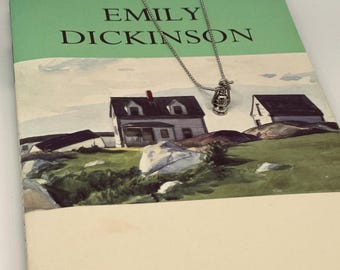 May include: A silver necklace with a small lantern charm, resting on a book titled "The Selected Poems of Emily Dickinson". The book cover features a watercolor painting of a white house with a green lawn.