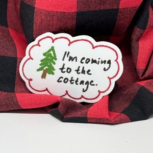 May include: A white, cloud-shaped sticker with a red border, featuring a green and brown tree illustration. The sticker has the handwritten text, "I'm coming to the cottage." The sticker rests on a red and black plaid fabric.