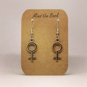 Può includere: Un paio di orecchini color argento con il simbolo femminile, appesi a ganci d'argento. Gli orecchini sono esposti su una scheda marrone con il testo "Mind the Book" stampato in alto.