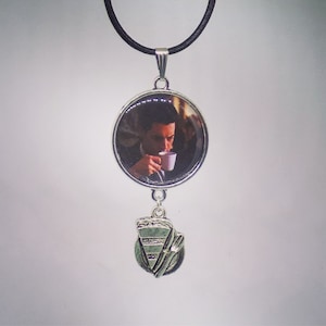May include: A silver pendant necklace with a circular image of a person drinking from a cup, and a pie slice charm. The necklace has a black cord and a silver bail. The pendant is suspended from a black cord.