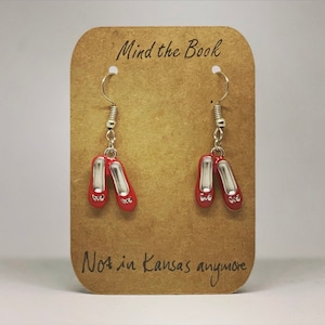 Può includere: Un paio di orecchini d'argento con ciondoli a forma di scarpette di rubino rosso. Gli orecchini sono esposti su una carta marrone con il testo "Mind the Book" e "Not in Kansas anymore".