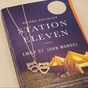 May include: A silver necklace with a gold and silver theatrical mask pendant. The necklace is lying on a book titled "Station Eleven" by Emily St. John Mandel.