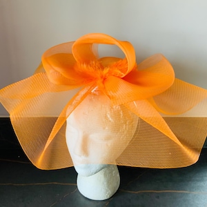 May include: A bright orange fascinator hat crafted from stiffened mesh netting. The headpiece features a large, structured bow design with wide, sweeping loops of sheer fabric that extend outwards, creating a voluminous and architectural silhouette.