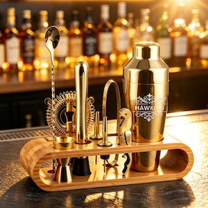 May include: A gold-coloured cocktail shaker set with a bamboo stand. The set includes a shaker with the text HAWKINS, a long mixing spoon, a jigger, a muddler, a strainer, a bottle opener, and pourers. All items are metallic gold and organised.