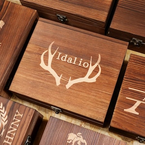 May include: A collection of rectangular dark brown wooden boxes with hinged lids and metal latches. Each box features unique laser-engraved designs, including the name Idalio centered between deer antlers, large monograms, and other custom text.