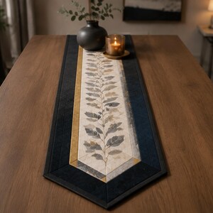May include: A rectangular table runner featuring a central cream-colored panel with a printed botanical leaf pattern in shades of gray and brown. The runner is bordered by thin strips of mustard yellow and wide bands of dark navy blue fabric.