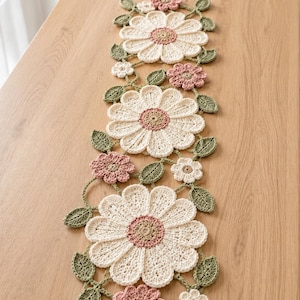 Può includere: Un runner da tavola rettangolare all'uncinetto con tre grandi motivi a margherita color crema con centri beige, intervallati da fiori più piccoli rosa polvere e bianchi, collegati da una rete di foglie e steli all'uncinetto verde salvia.