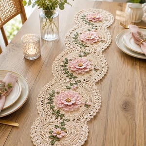 May include: A long, cream-colored crochet table runner featuring a series of connected circular motifs. Each circle is decorated with a three-dimensional, layered pink flower with a yellow center, accented by green crocheted vines and leaves.