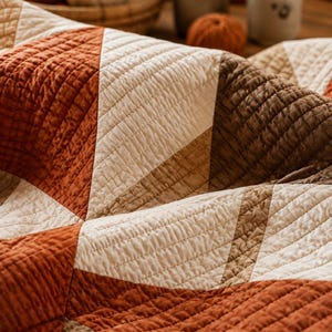 Pode incluir: Uma manta acolchoada com um padr&atilde;o geom&eacute;trico de tri&acirc;ngulos em tons de laranja ferrugem, creme e castanho escuro. O tecido apresenta uma textura de costura em grelha vis&iacute;vel em toda a superf&iacute;cie, criando um aspeto suave e em camadas.