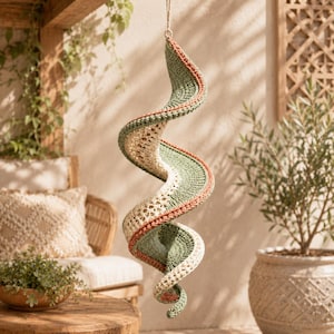 May include: A hanging spiral decoration crafted from crocheted yarn in alternating bands of sage green, cream, and terracotta orange. The structure features a continuous, twisting, ribbon-like form with a textured, open-stitch pattern throughout.