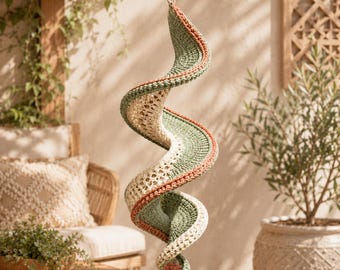 May include: A hanging spiral decoration crafted from crocheted yarn in alternating bands of sage green, cream, and terracotta orange. The structure features a continuous, twisting, ribbon-like form with a textured, open-stitch pattern throughout.