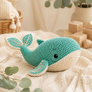 May include: A teal and cream-colored crocheted whale toy featuring a rounded body, a large tail fin with light pink stitched details, and a matching side fin. The whale has a smooth, textured surface, a small dorsal fin, and a black embroidered eye.