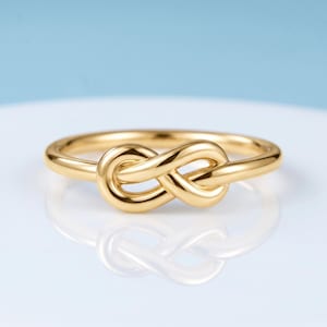 Peut inclure: Une bague en or poli présentant un motif de nœud infini central. L'anneau est lisse et circulaire, avec l'élément en forme de nœud centré sur le dessus. Le métal possède une finition brillante et réfléchissante qui capte la lumière.