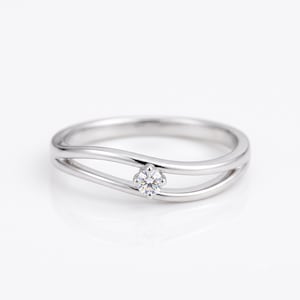 Puede incluir: Un anillo de tono plateado pulido con un diseño delicado de dos bandas fluidas que se curvan alrededor de una única piedra preciosa pequeña, redonda y transparente con facetas, fijada en el centro del espacio entre las dos bandas.
