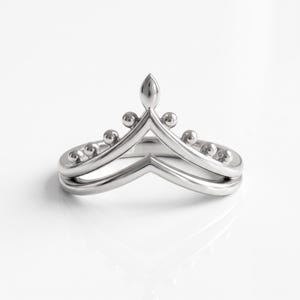 May include: A silver-toned metal ring featuring a double chevron or V-shaped band design. The upper band is adorned with a series of small, evenly spaced spherical beads and a central pointed element, creating a crown-like silhouette.