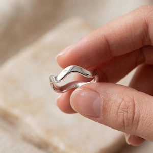 Op de afbeelding: Een ring van gepolijst zilver met een glad, doorlopend golvend ontwerp. De band heeft een reflecterend, metalen oppervlak met een enkele opwaartse bocht die een vloeiende, golvende vorm creëert. De ring is in een scherp beeld vastgelegd.