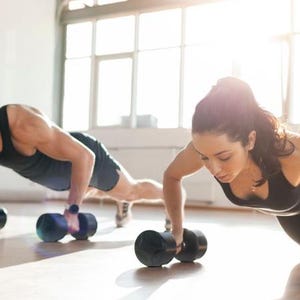 May include: Two individuals in black athletic tank tops and shorts performing push-ups while gripping black dumbbells. The exercise equipment features circular weights on each end of the handles, positioned on the floor during the workout.
