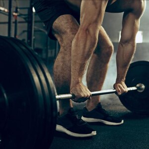 May include: A metal barbell with large black circular weight plates attached to each end. The barbell is positioned horizontally, held by a person in a gym setting, with the weight plates showing a smooth, solid black surface and a central metal hub.