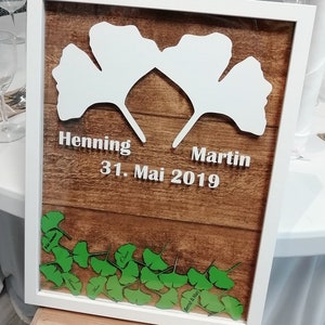 Könnte beinhalten: Ein weißer Holzrahmen mit braunem Holzgrund. Zwei weiße Ginkgoblätter sind an den Stielen verbunden. Die Blätter tragen die Namen Henning und Martin. Das Datum 31. Mai 2019 ist unter den Blättern geschrieben. Der untere Teil des Rahmens ist mit einer Reihe grüner Ginkgoblätter mit darauf geschriebenen Namen verziert.