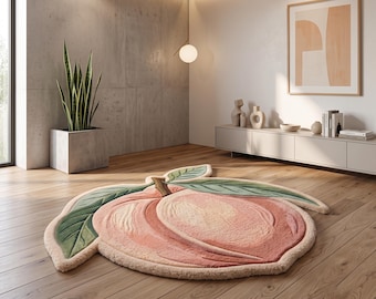 May include: A tufted area rug shaped like a peach, featuring a soft pink and cream gradient body with a brown stem and two large green leaves with detailed stitching. The rug has a thick, textured pile and is laid flat on a wooden floor.