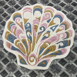 May include: A tufted rug shaped like a seashell with a white base and intricate, raised swirling patterns in shades of dusty pink, muted blue, and mustard yellow. The design features symmetrical, fan-like segments radiating from a central spiral point.