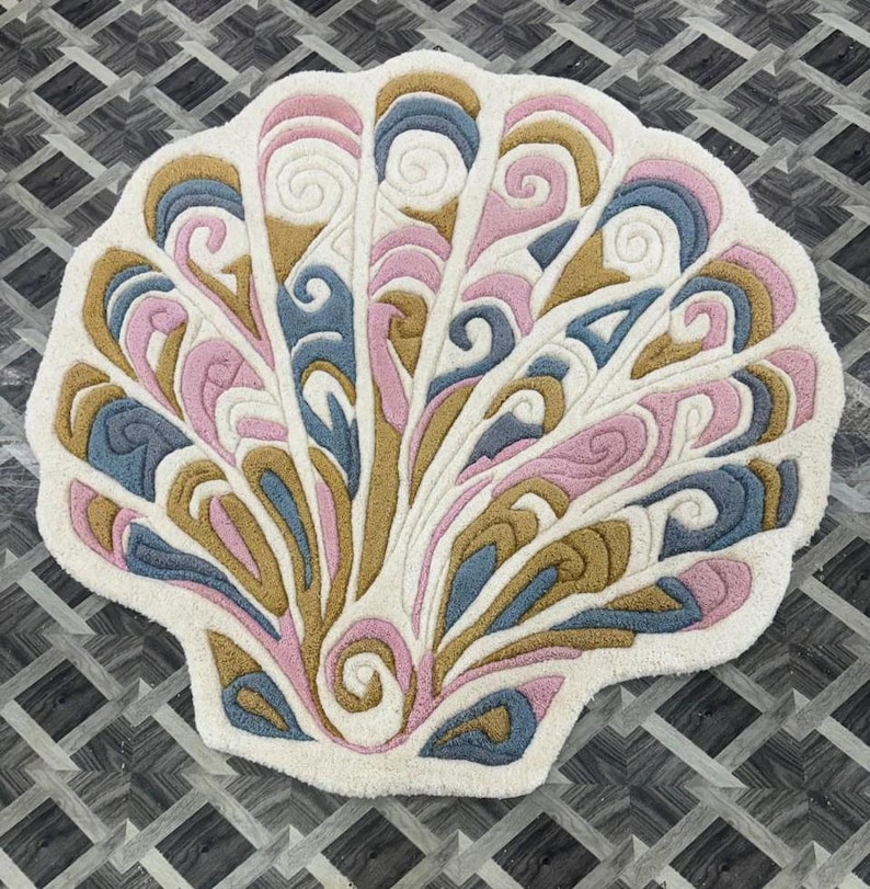 May include: A tufted rug shaped like a seashell with a white base and intricate, raised swirling patterns in shades of dusty pink, muted blue, and mustard yellow. The design features symmetrical, fan-like segments radiating from a central spiral point.