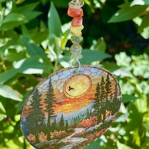 Può includere: Un acchiappasole circolare con una stampa paesaggistica in stile vetrata che raffigura un sentiero nel bosco, alti pini e una grande luna gialla con la sagoma di un uccello. L'ornamento pende da un filo di pietre irregolari, lucide e colorate.