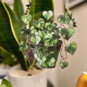 Può includere: Un acchiappasole in vetro colorato che raffigura una pianta in vaso con foglie verdi a forma di cuore e un vaso testurizzato. L'oggetto è realizzato con linee di saldatura in metallo scuro e presenta due fili di perline con accenti verdi.