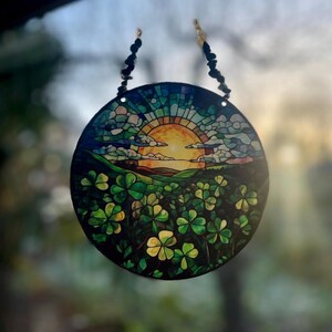 Può includere: Un acchiappasole circolare in vetro colorato raffigurante un sole giallo brillante che tramonta su un paesaggio verdeggiante pieno di trifogli. Il design include segmenti di cielo blu e viola ed è sospeso da due catenelle di perline.
