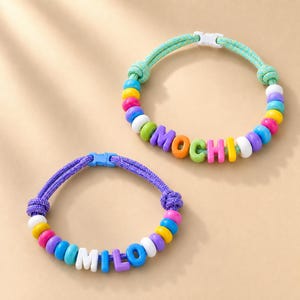 May include: Two adjustable bracelets made of woven cord with plastic side-release buckles. One bracelet features a light green cord with multicolored beads spelling MOCHI. The other bracelet has a purple cord with beads spelling MILO.