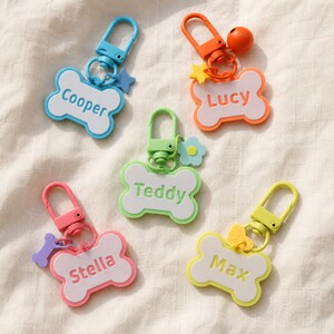 May include: Five bone-shaped keychains with colorful plastic clasps and charms. Each keychain features a white center with a colored border and a name printed in the middle: Cooper in blue, Lucy in orange, Teddy in green, Stella in pink, and Max in yellow.