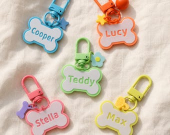 May include: Five bone-shaped keychains with colorful plastic clasps and charms. Each keychain features a white center with a colored border and a name printed in the middle: Cooper in blue, Lucy in orange, Teddy in green, Stella in pink, and Max in yellow.