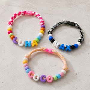 May include: Three bracelets featuring colorful cords and plastic letter beads. One bracelet has a pink and white cord with the name LUNA, one has a black and white patterned cord with the name MAX, and one has a peach cord with the name COCO.