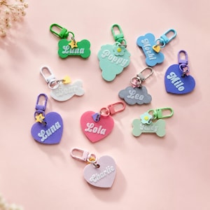 May include: A collection of colorful personalized keychains in various shapes including bones, hearts, and clouds. Each keychain features a name in white script, a small plastic charm, and a matching lobster clasp for attachment to items.