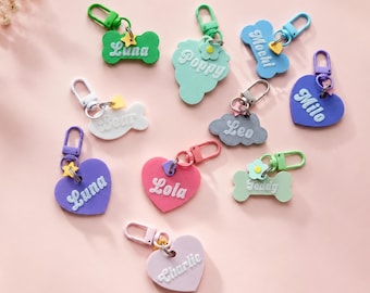 May include: A collection of colorful personalized keychains in various shapes including bones, hearts, and clouds. Each keychain features a name in white script, a small plastic charm, and a matching lobster clasp for attachment to items.