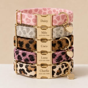 May include: A stack of five pet collars featuring various leopard print patterns in pink, gray, brown, and tan. Each collar has a gold-tone metal buckle engraved with a name and phone number, and a gold-tone heart-shaped charm attached to a D-ring.