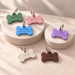 May include: Six bone-shaped pet ID tags in green, pink, blue, purple, cream, and brown. Each tag features a metal ring and engraved text including names like Finn, Bella, Milo, and Coco, or the phrase Call My Mom with a phone number.