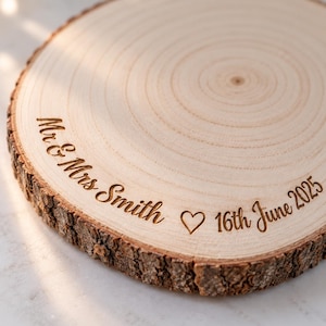 May include: A circular wooden slice with natural bark edges featuring laser-engraved text that reads Mr & Mrs Smith, a heart symbol, and the date 16th June 2025. The light-colored wood grain is visible across the flat surface of the round plaque.